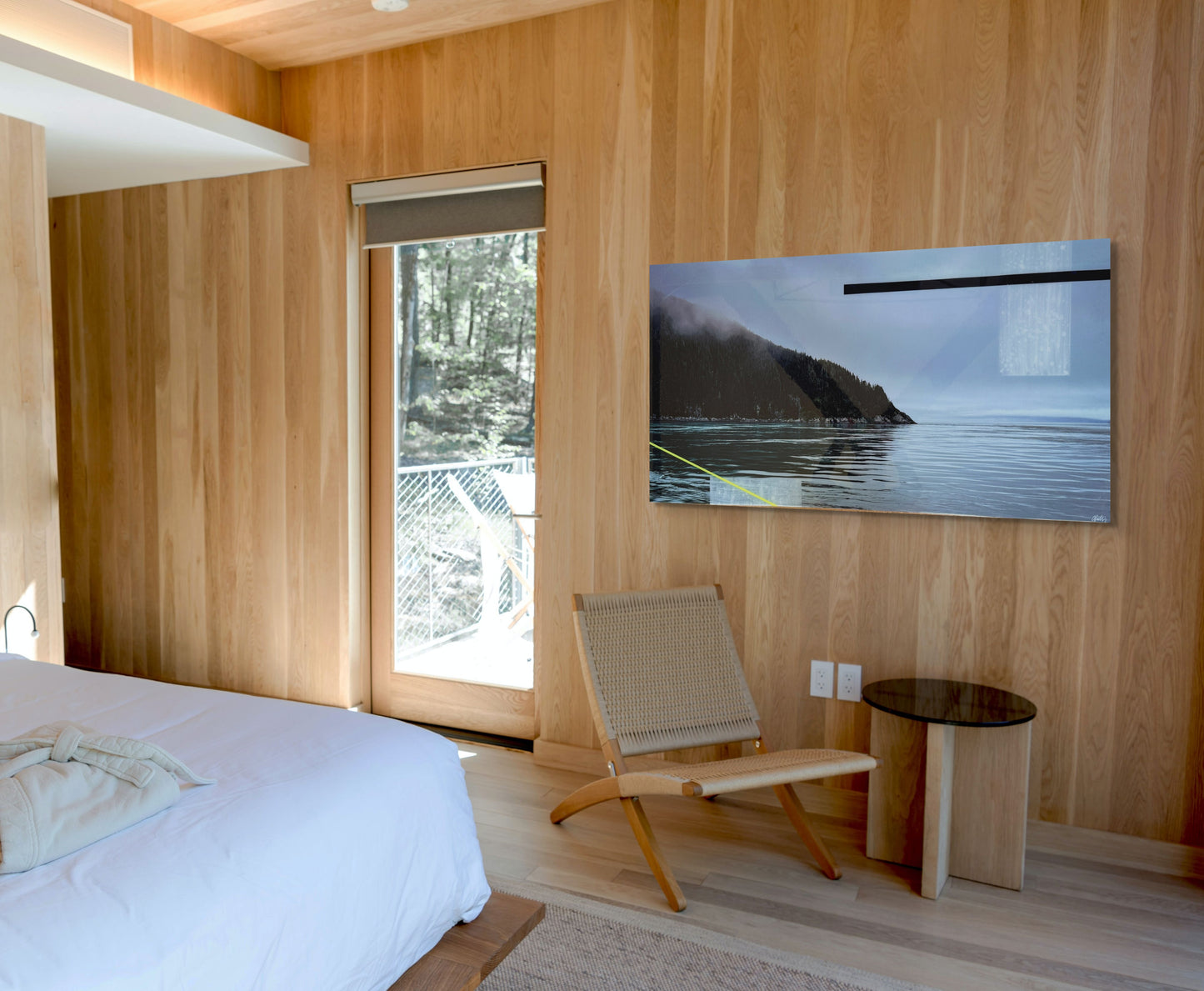 "'Alaskan Mist' by Christen Oatley displayed in a modern wood-paneled bedroom. The coastal landscape artwork adds serenity and depth above a woven lounge chair and side table, complementing the natural textures and minimalist design of the space.