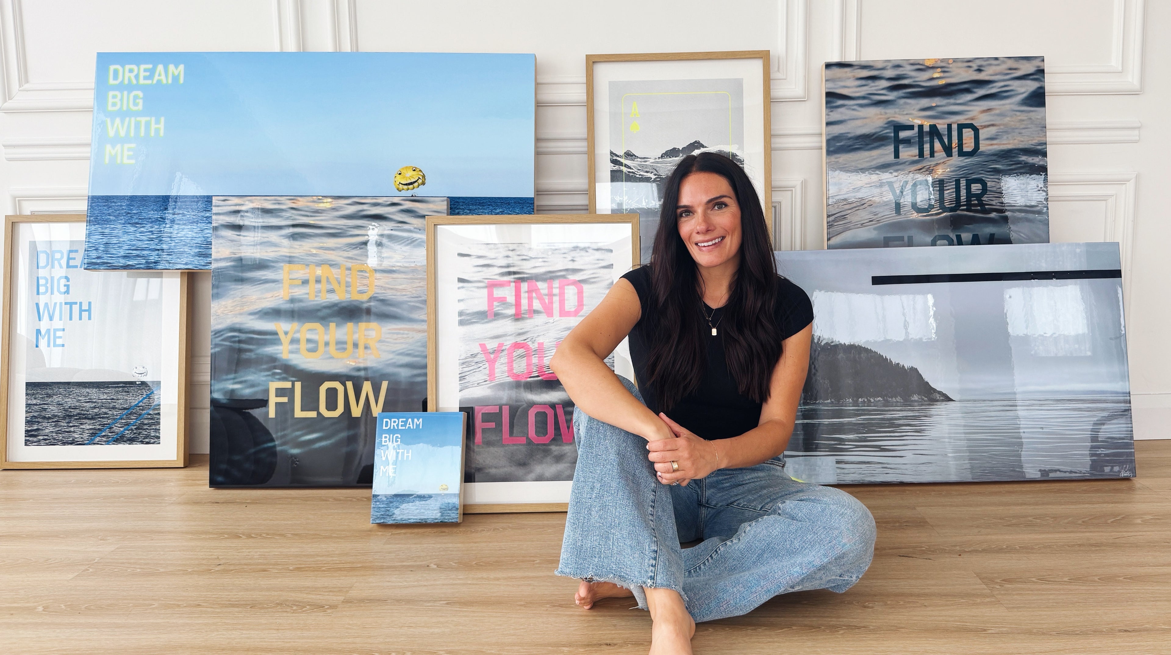Artist Christen Oatley sitting barefoot on a wooden floor, smiling in front of a collection of her framed ocean-inspired artwork. The pieces feature phrases like 'DREAM BIG WITH ME' and 'FIND YOUR FLOW' in bold lettering, set against serene water and nature scenes.