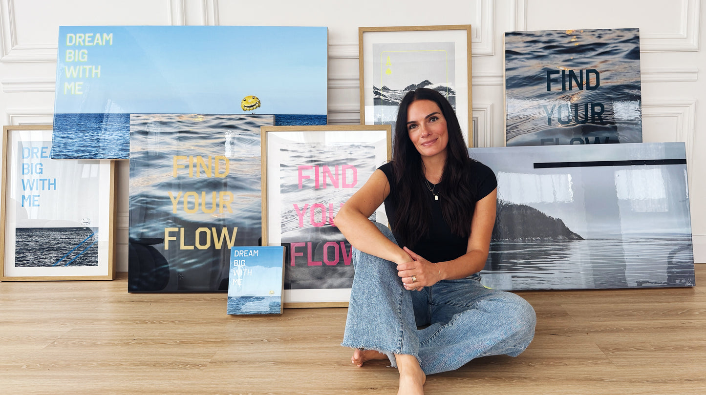 Artist Christen Oatley sitting barefoot on a wooden floor, smiling in front of a collection of her framed ocean-inspired artwork. The pieces feature phrases like 'DREAM BIG WITH ME' and 'FIND YOUR FLOW' in bold lettering, set against serene water and nature scenes.