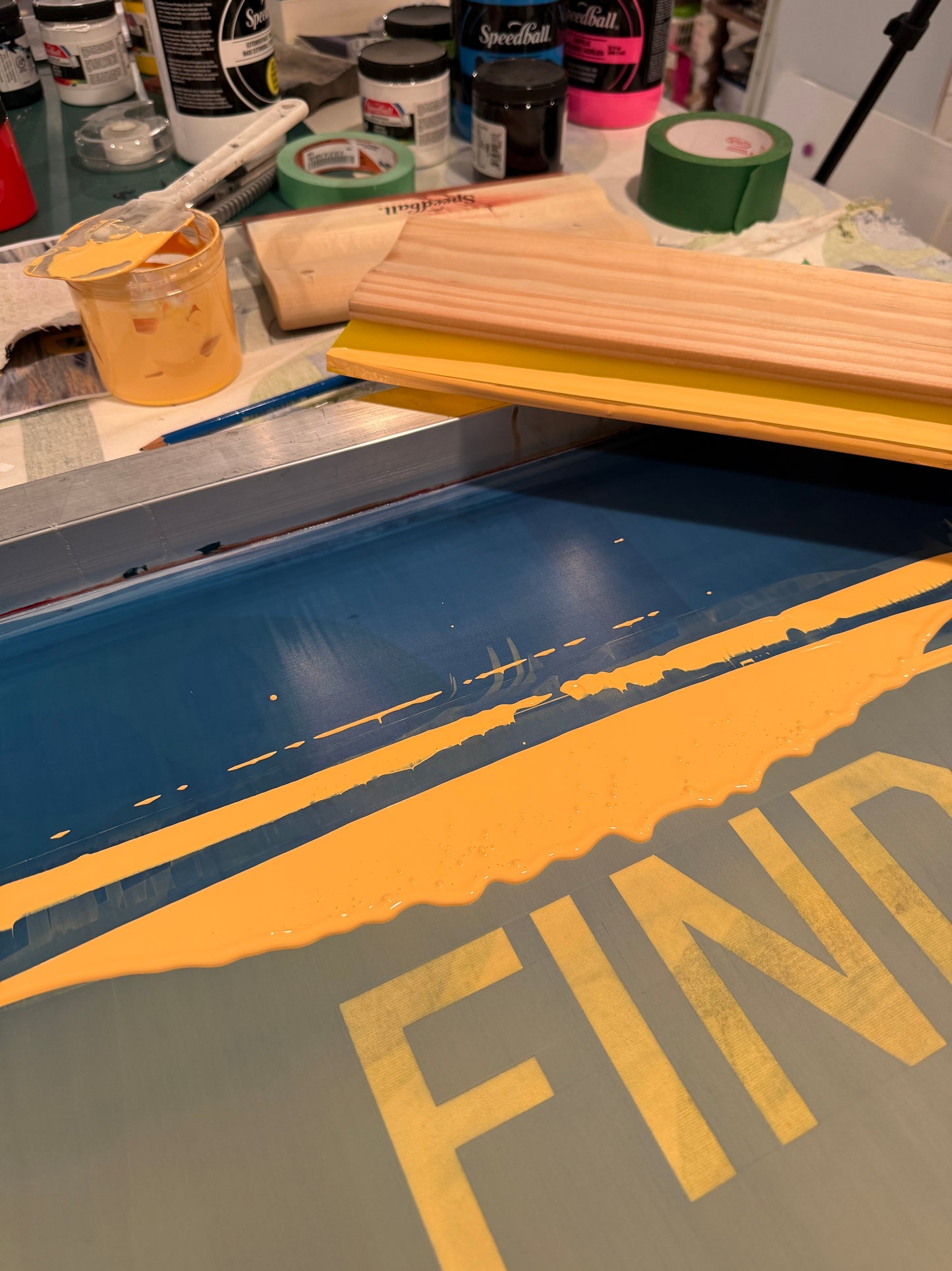 Behind the scenes of Christen Oatley's screen printing process for 'Find Your Flow – Sunset,' showing yellow acrylic ink being applied over a photographic print using a squeegee and stencil in a mixed media studio setup.