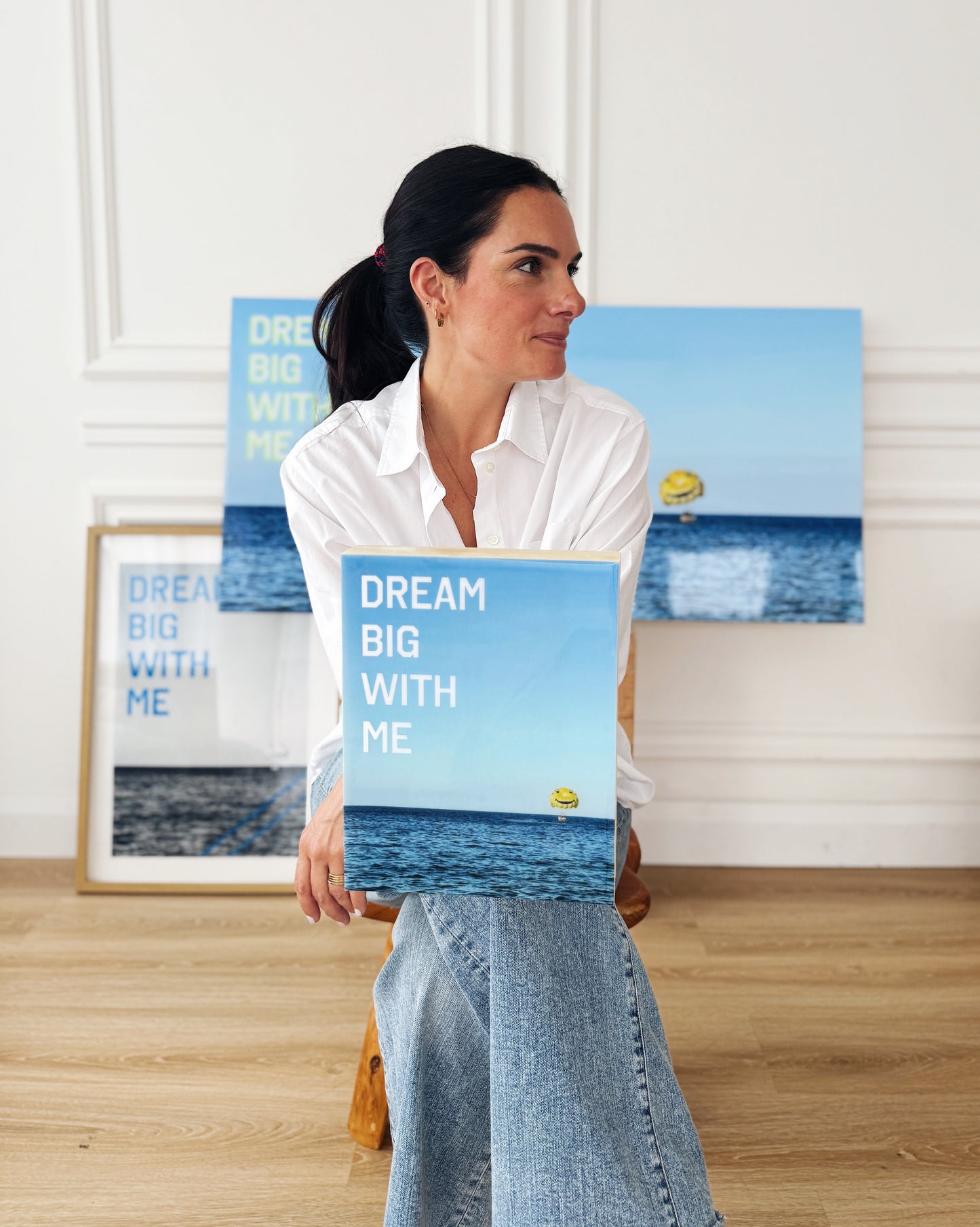 Artist Christen Oatley holding her limited edition mixed media piece 'Dream Big With Me' in a studio setting, surrounded by framed and resin-coated versions featuring a yellow parasail over the Mediterranean Sea.