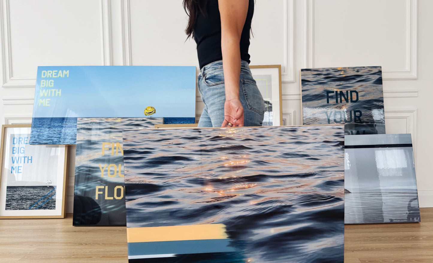 Artist Christen Oatley holding 'Sunset Tones' original mixed media artwork in a studio setting, surrounded by framed and resin-coated wall art featuring inspirational quotes and lake photography on birchwood panels.