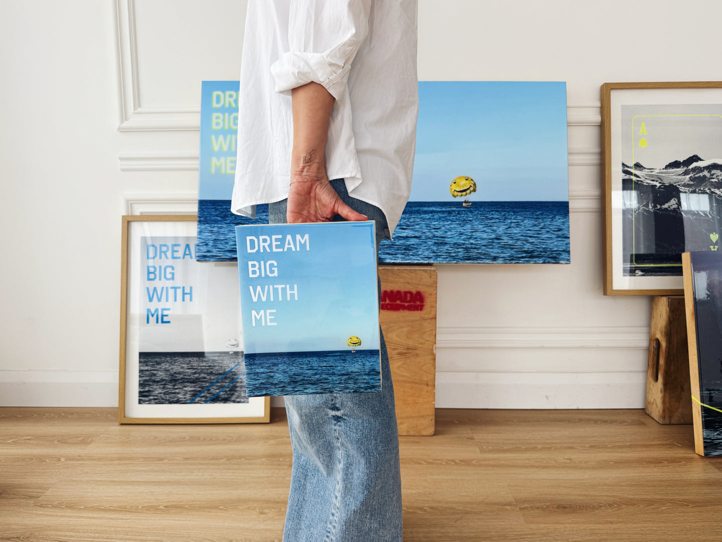 Artist Christen Oatley holding her 8x10 inch limited edition artwork 'Dream Big With Me,' surrounded by framed and resin-coated versions of the same parasail-over-sea photograph in a modern studio setting.
