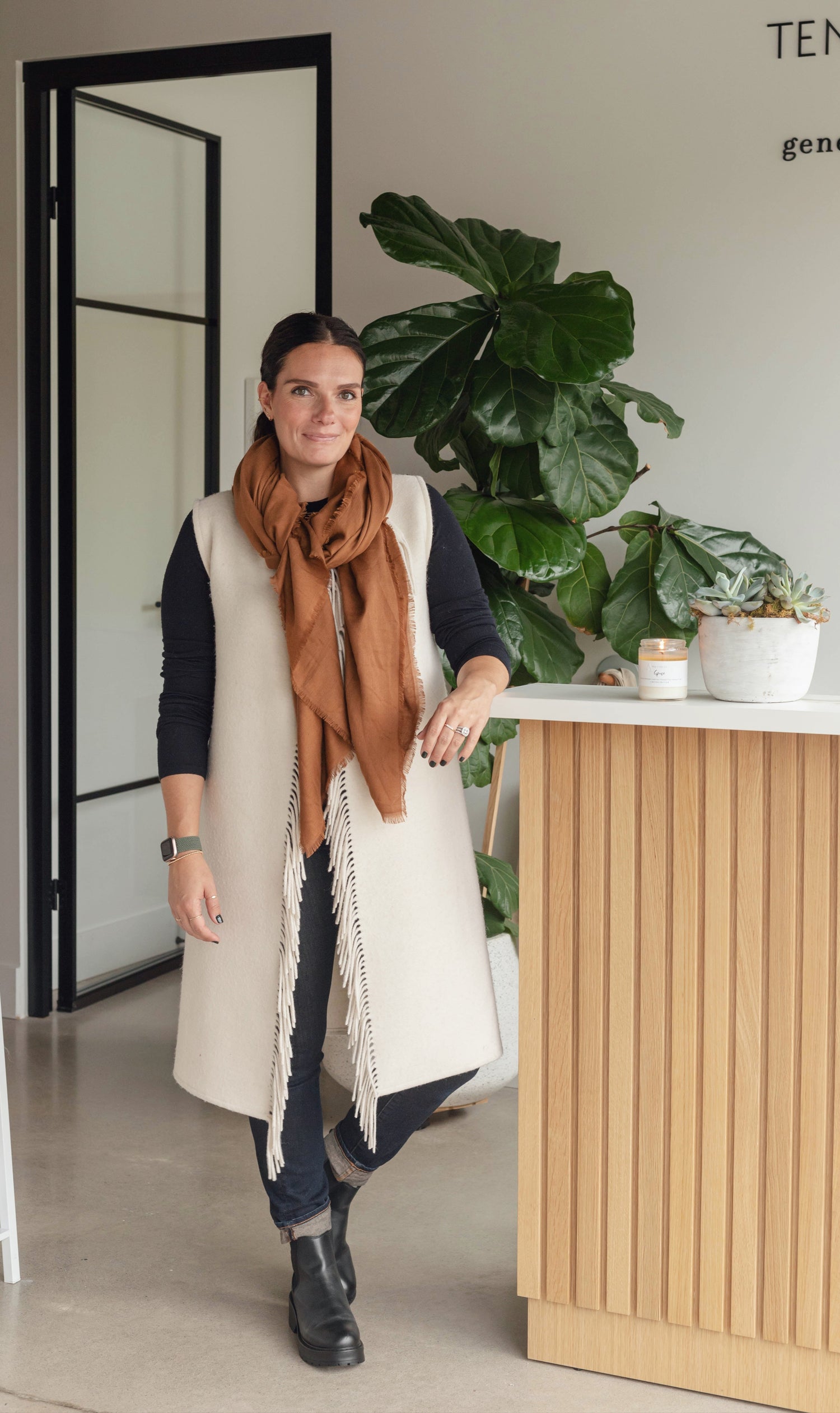 Artist Christen Oatley standing in a modern, minimalist space with neutral tones, wearing a long cream vest, rust-colored scarf, and black boots. She stands beside a wood-accented counter with potted plants and a candle, exuding a warm, approachable, and creative vibe.
