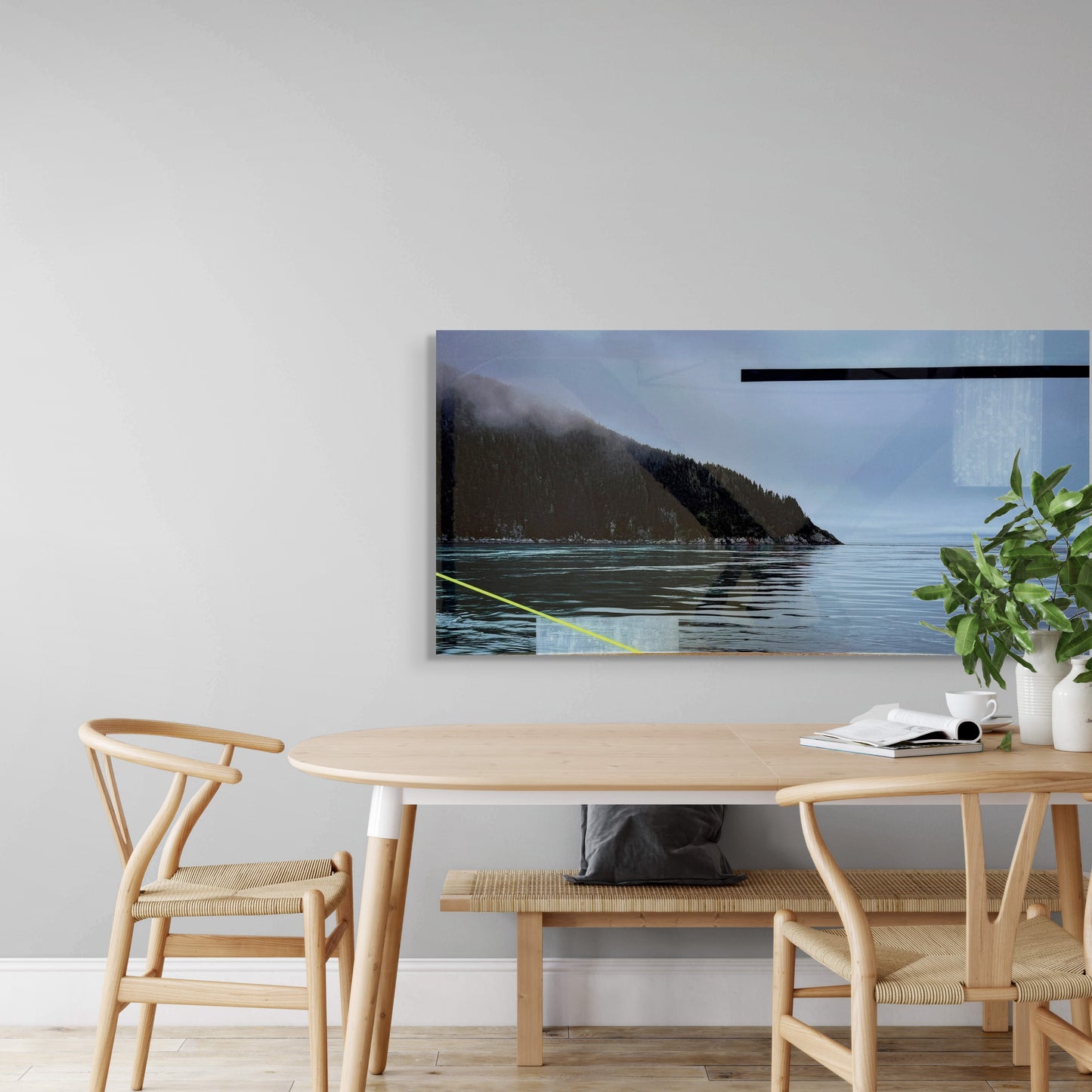 Modern dining area with a light wood table, two woven chairs, and a bench beneath a large framed coastal landscape artwork. The wall art features a misty forested cliffside beside calm ocean water, adding a tranquil and natural vibe to the minimalist space.