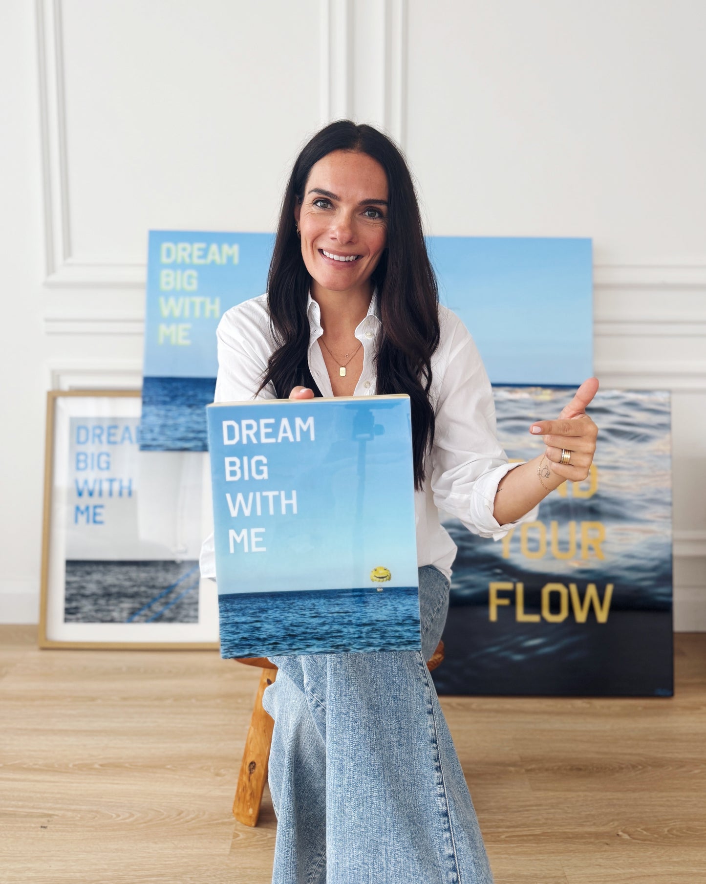 Artist Christen Oatley holding her limited edition mixed media artwork 'Dream Big With Me' in a studio setting, surrounded by additional resin-coated pieces including 'Find Your Flow' and framed photography prints.