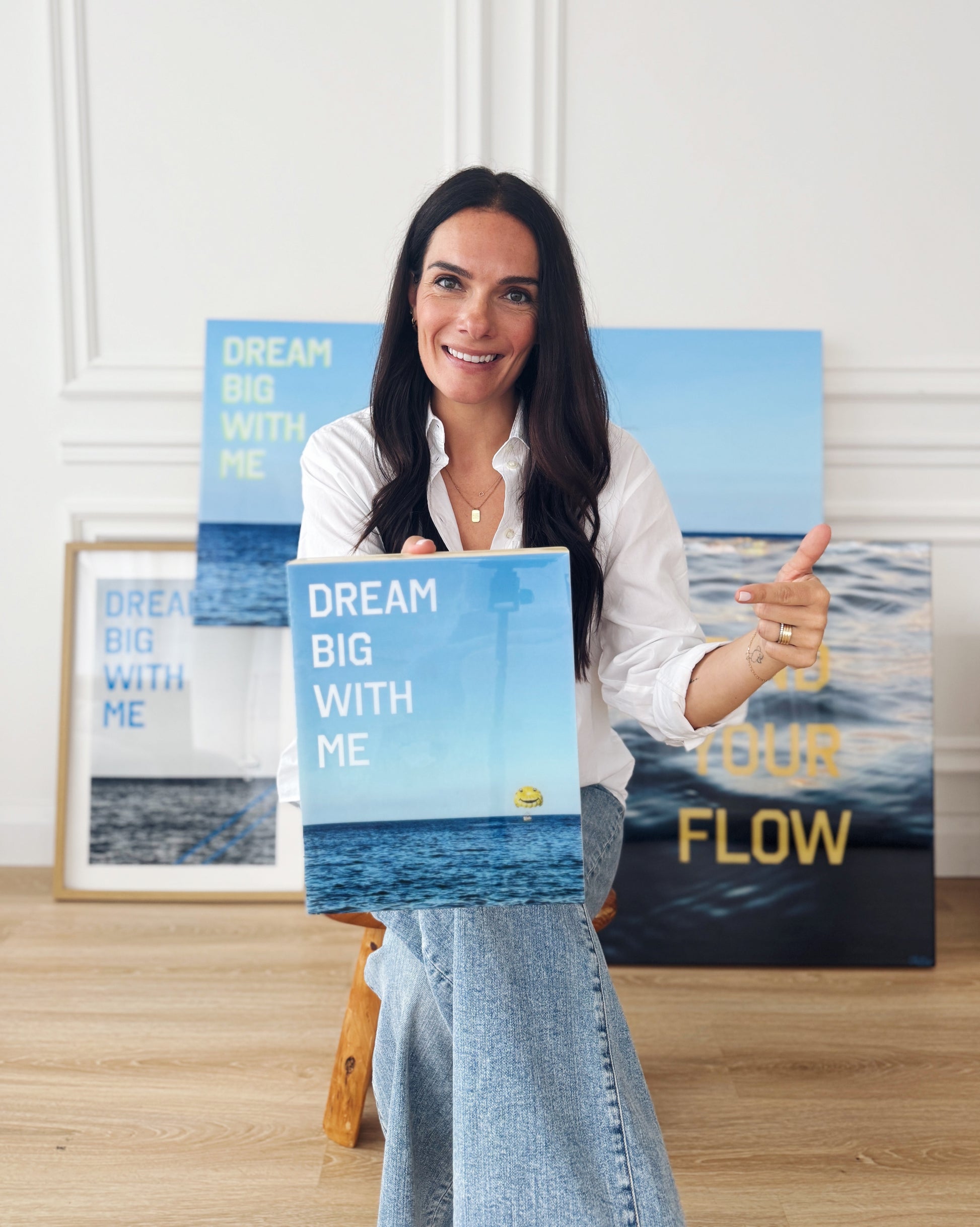 Artist Christen Oatley holding her limited edition mixed media artwork 'Dream Big With Me' in a studio setting, surrounded by additional resin-coated pieces including 'Find Your Flow' and framed photography prints.