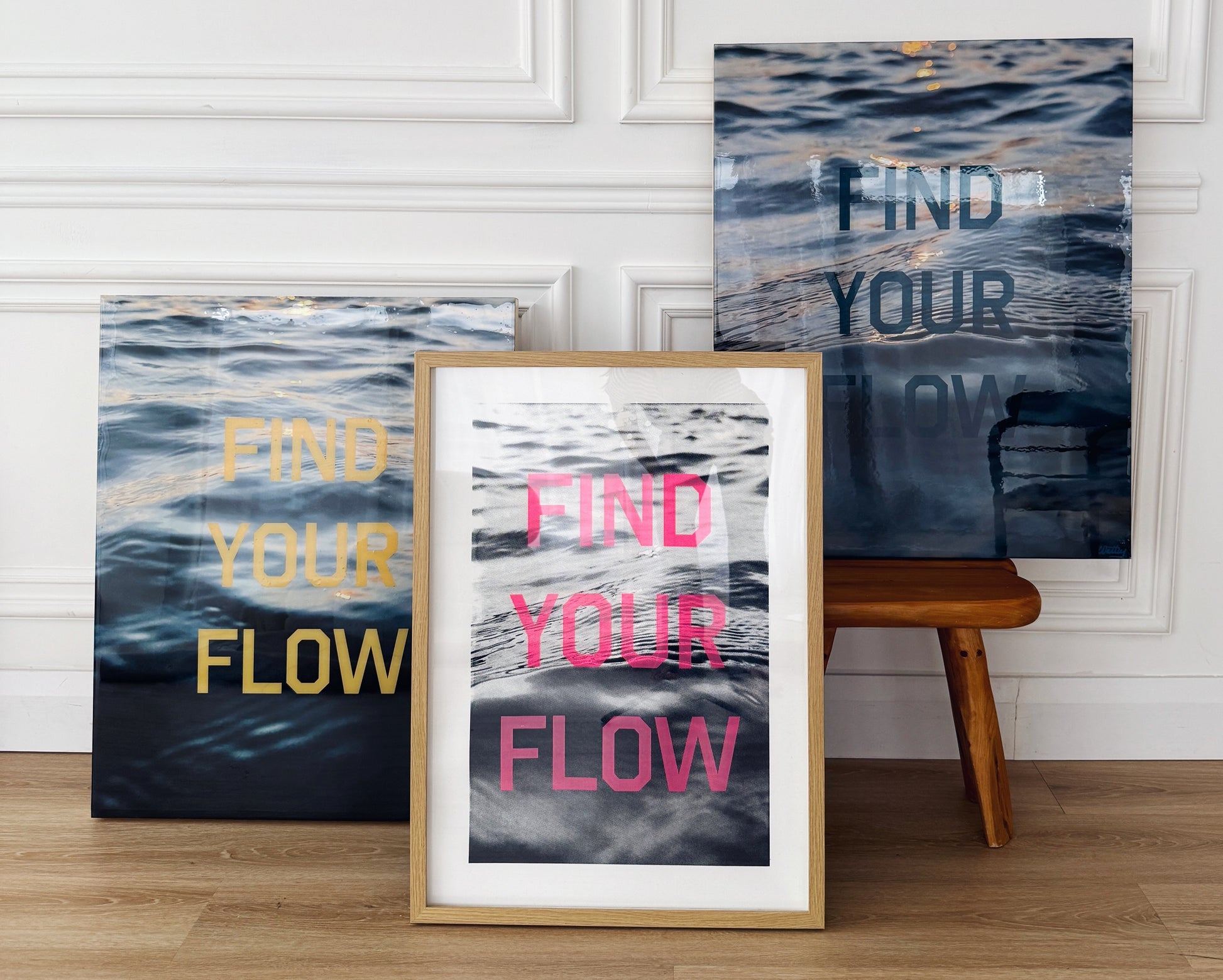 Three versions of Christen Oatley's 'Find Your Flow' artwork displayed together—original mixed media pieces with yellow and black screen-printed text on lake photography resin panels, and a framed pink text print on paper—showcasing artistic variations in a modern studio setting.